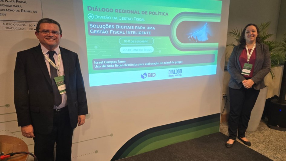 A foto mostra a subsecretária do Tesouro do Estado, Juliana Debaquer, e o gerente do projeto de precificação, Israel Campos Fama, posicionados em frente a uma tela de projeção. Ambos usam crachás de identificação e trajes formais. Ao fundo, o painel destaca o evento “Diálogo Regional de Política – Soluções Digitais para uma Gestão Fiscal Inteligente”, promovido pelo Banco Interamericano de Desenvolvimento (BID).