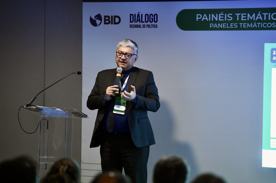 A imagem mostra o subsecretário da Receita Estadual, Ricardo Neves Pereira, em apresentação durante o evento do Banco Interamericano de Desenvolvimento (BID). Ele fala ao microfone enquanto gesticula, em frente a um telão com os dizeres “Painéis Temáticos – Diálogo Regional de Política”. Usa traje social e crachá de identificação.