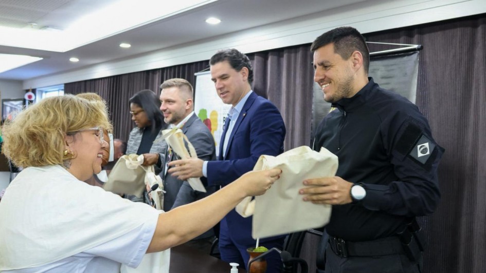 Foto em ambiente fechado de uma mulher, de roupa branca, entregando uma ecobag branca a um participante do evento, de roupas escuras, no primeiro plano. Ambos sorriem e estão um de frente para o outro, e de lado para a câmera. Ao fundo, aparecem dois homens e uma mulher de lado, também com ecobags.