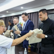 Foto em ambiente fechado de uma mulher, de roupa branca, entregando uma ecobag branca a um participante do evento, de roupas escuras, no primeiro plano. Ambos sorriem e estão um de frente para o outro, e de lado para a câmera. Ao fundo, aparecem dois homens e uma mulher de lado, também com ecobags.