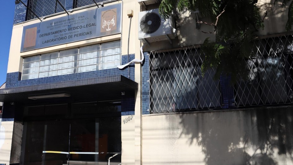 A imagem mostra a fachada de um prédio público com revestimento em pastilhas azuis. No centro superior da fachada há uma placa metálica com os seguintes dizeres:
ESTADO DO RIO GRANDE DO SUL
Secretaria da Segurança Pública
INSTITUTO-GERAL DE PERÍCIAS
Departamento Médico-Legal
Laboratório de Perícias
Acima da placa, há o brasão do Instituto-Geral de Perícias (IGP-RS) e outro símbolo institucional. À direita da entrada, há aparelhos de ar-condicionado instalados na parede. A entrada principal do prédio possui portas de vidro e uma pequena escada com corrimão metálico. À esquerda da entrada está o número 1807, indicando o endereço. As janelas do prédio possuem grades de proteção em padrão cruzado. Uma árvore projeta sombra sobre parte da fachada.