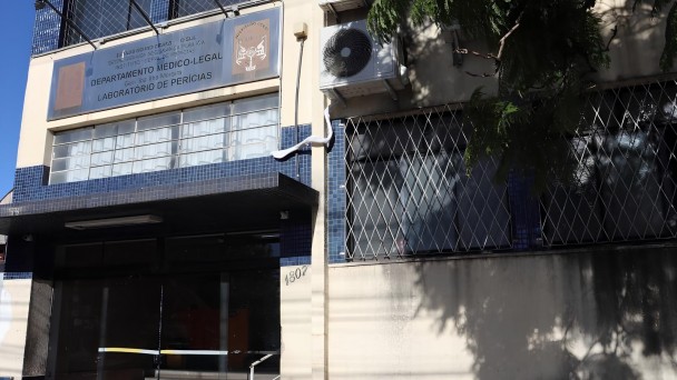 A imagem mostra a fachada de um prédio público com revestimento em pastilhas azuis. No centro superior da fachada há uma placa metálica com os seguintes dizeres:

ESTADO DO RIO GRANDE DO SUL
Secretaria da Segurança Pública
INSTITUTO-GERAL DE PERÍCIAS
Departamento Médico-Legal
Laboratório de Perícias

Acima da placa, há o brasão do Instituto-Geral de Perícias (IGP-RS) e outro símbolo institucional. À direita da entrada, há aparelhos de ar-condicionado instalados na parede. A entrada principal do prédio possui portas de vidro e uma pequena escada com corrimão metálico. À esquerda da entrada está o número 1807, indicando o endereço. As janelas do prédio possuem grades de proteção em padrão cruzado. Uma árvore projeta sombra sobre parte da fachada.
