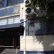 A imagem mostra a fachada de um prédio público com revestimento em pastilhas azuis. No centro superior da fachada há uma placa metálica com os seguintes dizeres:

ESTADO DO RIO GRANDE DO SUL
Secretaria da Segurança Pública
INSTITUTO-GERAL DE PERÍCIAS
Departamento Médico-Legal
Laboratório de Perícias

Acima da placa, há o brasão do Instituto-Geral de Perícias (IGP-RS) e outro símbolo institucional. À direita da entrada, há aparelhos de ar-condicionado instalados na parede. A entrada principal do prédio possui portas de vidro e uma pequena escada com corrimão metálico. À esquerda da entrada está o número 1807, indicando o endereço. As janelas do prédio possuem grades de proteção em padrão cruzado. Uma árvore projeta sombra sobre parte da fachada.