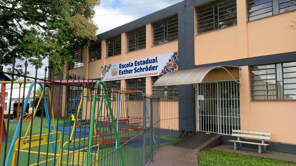 A imagem mostra a fachada da Escola Estadual Esther Schröder, um prédio de dois andares com paredes em tons de bege e cinza, localizado atrás de um pátio com brinquedos coloridos, como balanços e escorregadores, indicativos de um espaço voltado para crianças.
Acima da entrada principal da escola há uma grande faixa com o nome completo da instituição e o lema:
"Educação que acolhe, valores que transformam."
A entrada é protegida por um portão de grade branca com uma pequena cobertura metálica curva. À frente do prédio há uma cerca de ferro e uma calçada que leva até uma rampa de acessibilidade. Também há um pequeno banco pintado de branco e azul, posicionado ao lado da entrada. Uma árvore grande e frondosa está visível à esquerda, projetando sombra sobre o pátio.
O ambiente parece limpo, organizado e acolhedor, transmitindo a sensação de um espaço seguro e dedicado à educação infantil e fundamental.