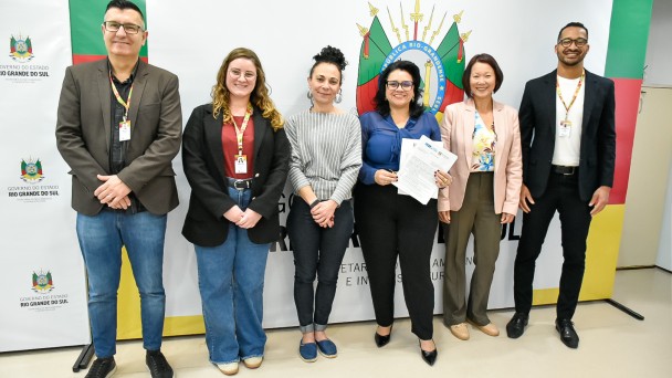 Seis pessoas estão alinhadas em pé, posando para a foto diante de um painel institucional do Governo do Estado do Rio Grande do Sul, que traz o brasão oficial no centro e as cores verde, vermelho e amarelo nas laterais. Da esquerda para a direita:

Um homem de óculos, cabelo curto escuro, veste blazer marrom, camisa preta e jeans.

Uma mulher de cabelos ondulados loiros, óculos e sorriso, veste blazer preto, blusa vinho e calça jeans larga.

Uma mulher de cabelos presos em coque alto, veste blusa cinza listrada e calça preta.

Uma mulher de cabelos escuros soltos, óculos, camisa azul e calça preta, segura papéis nas mãos.

Uma mulher de cabelos curtos castanhos, veste blazer rosa-claro, blusa estampada e calça marrom.

Um homem de barba curta, óculos, blazer preto, camiseta branca e calça escura.

Todos estão sorrindo e usam crachás de identificação, sugerindo um ambiente institucional ou oficial.