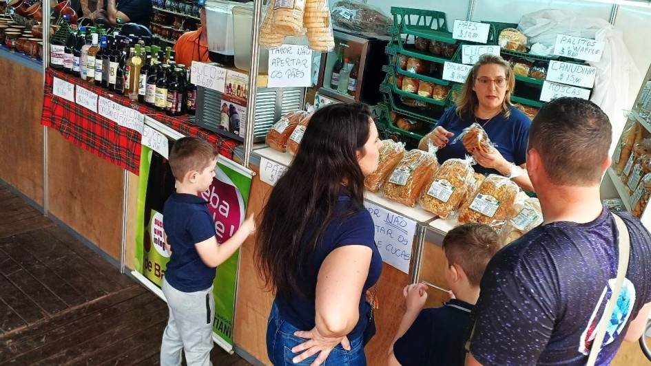 A imagem mostra uma cena movimentada de feira em um estande de venda de pães e produtos coloniais. No centro da imagem, uma mulher loira, vestida com camiseta azul, está atendendo uma família composta por dois adultos e duas crianças. Ela segura um pacote de pão e parece estar explicando algo sobre o produto. Sobre o balcão estão expostos diversos pacotes de pães, cucas e produtos similares, embalados em plástico transparente.
Ao fundo, prateleiras verdes estão cheias de mais pães e bolachas. Placas escritas à mão informam os preços dos produtos, como “PÃES $10,00”, “CUCA”, “BOLACHA $10,00”, “CANUDINHO $15,00”, e “PALITOS WAFFLE $8,00”. Outra placa no balcão diz “POR FAVOR NÃO APERTAR OS PÃES E CUCAS”.
À esquerda, outro estande vende vinhos, sucos e produtos artesanais, com garrafas expostas e utensílios de cerâmica pendurados. Há uma faixa vermelha xadrez decorando a banca. Crianças observam curiosas os produtos enquanto os pais conversam com a vendedora.
O ambiente é rústico, com piso de madeira, típico de feiras coloniais ou de produtos artesanais. A atmosfera transmite um clima familiar e acolhedor.