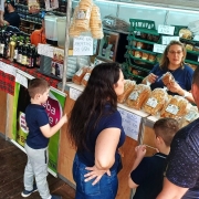 A imagem mostra uma cena movimentada de feira em um estande de venda de pães e produtos coloniais. No centro da imagem, uma mulher loira, vestida com camiseta azul, está atendendo uma família composta por dois adultos e duas crianças. Ela segura um pacote de pão e parece estar explicando algo sobre o produto. Sobre o balcão estão expostos diversos pacotes de pães, cucas e produtos similares, embalados em plástico transparente.

Ao fundo, prateleiras verdes estão cheias de mais pães e bolachas. Placas escritas à mão informam os preços dos produtos, como “PÃES $10,00”, “CUCA”, “BOLACHA $10,00”, “CANUDINHO $15,00”, e “PALITOS WAFFLE $8,00”. Outra placa no balcão diz “POR FAVOR NÃO APERTAR OS PÃES E CUCAS”.

À esquerda, outro estande vende vinhos, sucos e produtos artesanais, com garrafas expostas e utensílios de cerâmica pendurados. Há uma faixa vermelha xadrez decorando a banca. Crianças observam curiosas os produtos enquanto os pais conversam com a vendedora.

O ambiente é rústico, com piso de madeira, típico de feiras coloniais ou de produtos artesanais. A atmosfera transmite um clima familiar e acolhedor.