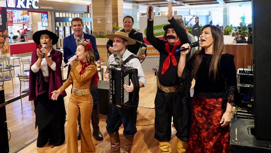 Cena de apresentação musical em shopping. Os artistas estão alinhados, todos com microfones, exceto um jovem com acordeão. Ao fundo, aparece a fachada de uma lanchonete. As roupas são típicas gaúchas, com bombachas, chapéus, lenços vermelhos e vestidos longos. Um dos artistas ergue os braços em gesto de animação.