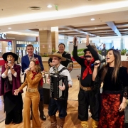 Cena de apresentação musical em shopping. Os artistas estão alinhados, todos com microfones, exceto um jovem com acordeão. Ao fundo, aparece a fachada de uma lanchonete. As roupas são típicas gaúchas, com bombachas, chapéus, lenços vermelhos e vestidos longos. Um dos artistas ergue os braços em gesto de animação.