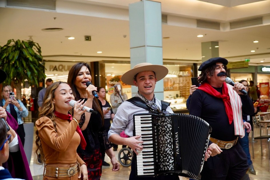 Quatro artistas em primeiro plano realizam apresentação: a cantora Luiza Barbosa, uma jovem com lenço vermelho canta, ao lado da cantora Cristina Sorrentino, uma mulher de vestido preto e vermelho também com microfone. Thomas Machado toca acordeão usando chapéu típico e, ao lado, o artista Jair Kobe caracterizado como Guri de Uruguaiana segura microfone. Atrás, pessoas assistem e registram com celulares.