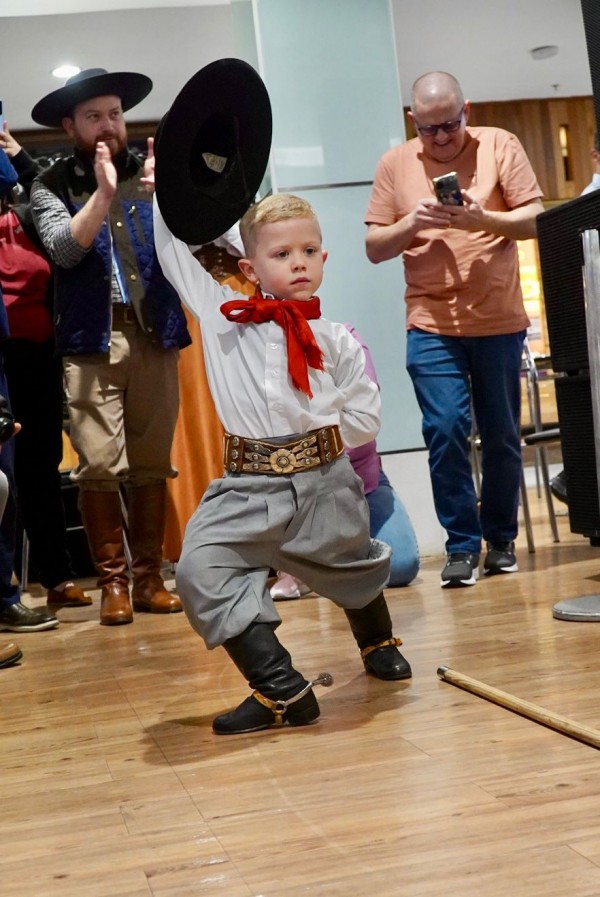 Um menino vestido com traje típico gaúcho dança em primeiro plano, erguendo o chapéu preto com uma mão e mantendo a outra na cintura. Ele usa bombacha cinza, camisa branca, lenço vermelho e botas pretas. Ao fundo, adultos assistem, fotografam e batem palmas.