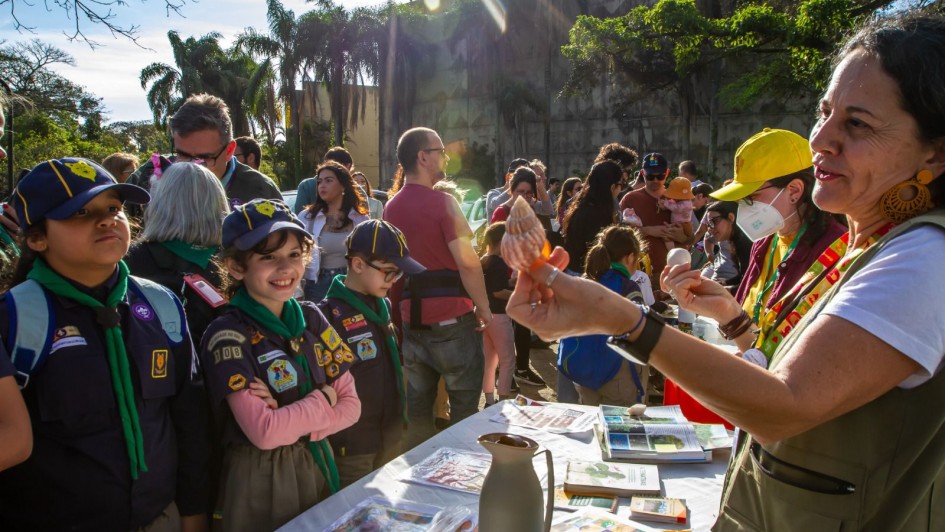 A imagem mostra um evento ao ar livre com crianças escoteiras, vestindo uniformes azuis com lenços verdes, observando uma mulher que segura e explica algo. Há mesas com livros e materiais educativos, além de várias pessoas ao fundo.