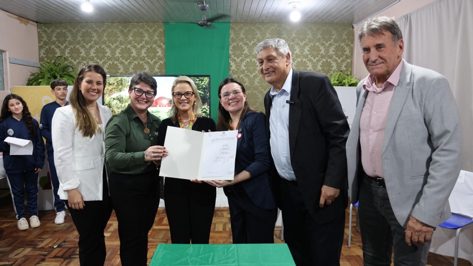 Foto em uma sala pintada em tons de verde. Quatro mulheres e dois homens posam para a foto. As duas mulheres ao centro seguram um documento assinado. Ao fundo, aparecem dois estudantes com uniformes na cor azul marinho. 