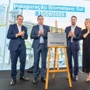 Fotografia da cerimônia de inauguração da Biometano Sul, realizada em 15 de setembro de 2025, em Minas do Leão (RS). No centro da imagem, há uma placa metálica sobre cavalete de madeira, com os dizeres “Unidade de Geração de Biometano em Minas do Leão – inaugurada em 15 de setembro de 2025 – Biometano Sul”. Ao redor da placa, quatro pessoas em trajes formais posam para a foto: três homens de terno e uma mulher de vestido preto, todos sorrindo e aplaudindo. Ao fundo, um telão exibe o título “Inauguração Biometano Sul – 15/09/2025”.