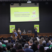 Na imagem, um auditório lotado de pessoas acompanha uma apresentação. No palco, há três cadeiras, sendo duas ocupadas por palestrantes sentados e uma terceira vazia. À direita, uma pessoa fala em pé com microfone em mãos. À esquerda, uma intérprete de Libras traduz a apresentação. Ao fundo, um grande telão exibe a frase “Ensino Médio em Tempo Integral – Fórum de Protagonismo, Chimas e Prosa”, acompanhado de logotipos institucionais. Também há dois banners verdes com o slogan “O futuro hoje” posicionados ao lado do palco.