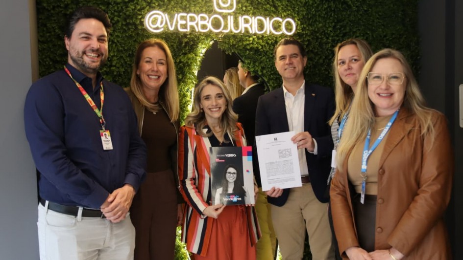 Seis pessoas estão posando para uma foto em um ambiente interno, diante de uma parede verde de plantas artificiais com o letreiro iluminado “@VERBOJURIDICO”. No centro, uma mulher de cabelos loiros e lisos veste blazer listrado em vermelho, azul e branco e segura uma revista com a capa exibindo sua própria foto. Ao lado dela, um homem de blazer azul e camisa branca segura um documento impresso. Os demais, três mulheres e um homem, estão sorridentes, vestidos com roupas sociais em tons de marrom, azul e bege, compondo uma atmosfera formal e cordial de registro institucional.
