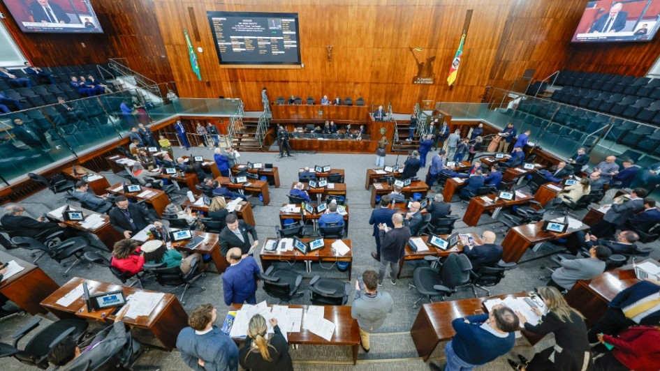 Fotografia mostra o plenário da Assembleia Legislativa do Rio Grande do Sul durante sessão. Deputados e deputadas ocupam seus lugares em bancadas de madeira dispostas em semicírculo. Algumas pessoas estão em pé, outras sentadas, manuseando documentos ou conversando. No fundo, sobre a parede de madeira, há o brasão do Estado e a bandeira do Rio Grande do Sul, ao lado da bandeira do Brasil. Um grande painel eletrônico exibe informações da sessão, enquanto telões nas laterais projetam imagens do presidente da mesa diretora.