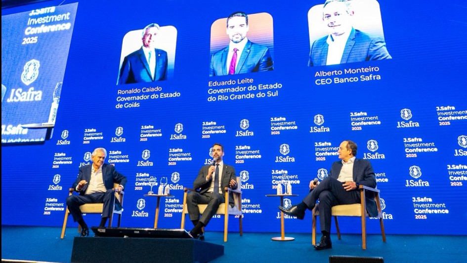 Três homens de terno estão sentados em poltronas no palco de um evento, com um telão azul ao fundo que mostra os nomes e fotos deles. O painel se chama “Desafios para o Brasil”. Leite está no meio.