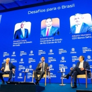 Três homens de terno estão sentados em poltronas no palco de um evento, com um telão azul ao fundo que mostra os nomes e fotos deles. O painel se chama “Desafios para o Brasil”. Leite está no meio.