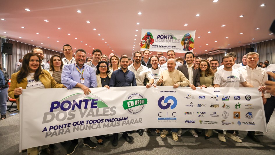 Um grupo numeroso de pessoas posa sorridente em um auditório, segurando uma grande faixa branca com a inscrição em destaque: “Ponte dos Vales – A ponte que todos precisam para nunca mais parar”. No centro da faixa aparecem os logotipos do movimento liderado pela CIC Vale do Taquari e de diversas entidades apoiadoras, como FIERGS, Sistema Farsul, Fecomércio RS e Federasul, entre outras. Algumas pessoas vestem camisetas brancas com o mesmo slogan da campanha. Ao fundo, outra faixa pendurada reforça a identidade do movimento, decorada com a imagem de uma ponte e uma bandeira do Rio Grande do Sul estilizada. O ambiente é iluminado com luzes de teto, e todos aparentam estar em clima de união e apoio ao projeto.