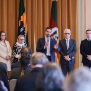 Cinco pessoas em pé diante de uma platéia; a do centro fala ao microfone. Atrás, bandeiras do Brasil e outra com vermelho, verde e branco. Ambiente formal, tipo conferência.

