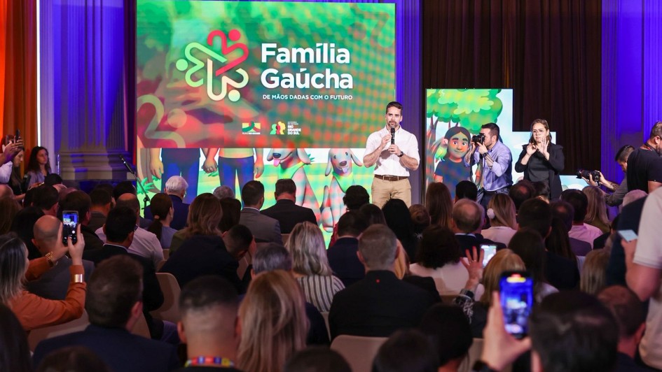 O governador Eduardo Leite está em pé no palco, falando ao microfone diante de uma plateia cheia. Ele veste camisa branca e calça bege. Ao fundo, um telão exibe a identidade visual do Programa Família Gaúcha, com o slogan “De mãos dadas com o futuro”.