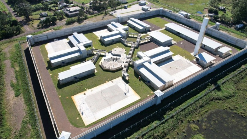 Visão aérea de uma instalação, possivelmente uma prisão ou complexo de segurança. O layout apresenta várias construções interligadas por passarelas, com cercas altas ao redor. Também podemos ver uma área de esporte ao fundo, provavelmente destinada à recreação dos internos. O design parece organizado e separado em várias seções, com estruturas de controle de segurança visíveis, como torres e muros. Há um espaço aberto com grama e alguns prédios adicionais ao fundo, sugerindo uma área ampla.