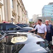 O governador Eduardo Leite aparece em frente ao Palácio Piratini, acompanhado de outras autoridades e assessores. Ele veste camisa branca e calça bege, caminhando ao lado de viaturas da Polícia Civil estacionadas na rua. Há também imprensa registrando o momento.