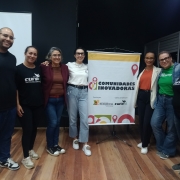 A imagem mostra um grupo de sete pessoas posando para uma foto em frente a um banner com o nome "COMUNIDADES INOVADORAS". O banner também inclui logos de organizações apoiadoras. As pessoas estão sorrindo e se vestem de forma casual, com camisetas e roupas variadas. O evento parece estar relacionado ao empreendedorismo ou qualificação, dado o contexto do banner e o ambiente de encontro. O fundo é de um espaço fechado, com piso de madeira e iluminação suave.