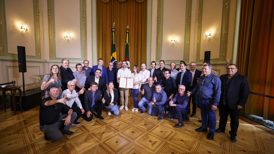 Um grupo de cerca de 30 pessoas posa para foto oficial em um salão nobre, com cortinas douradas e duas bandeiras ao fundo — a do Brasil e a do Rio Grande do Sul. No centro da imagem, duas pessoas seguram juntas um documento aberto, exibido para a câmera. Todos sorriem, alguns fazem sinal de positivo com as mãos. As pessoas estão dispostas em pé e agachadas na frente, em trajes formais ou sociais, indicando um evento oficial em ambiente governamental. O piso é de madeira em padrão geométrico e o teto tem iluminação com lustres de parede.