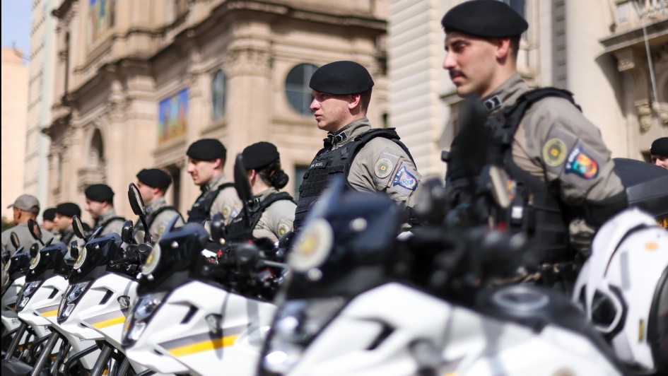 Policiais militares da Brigada Militar estão perfilados em frente a uma fileira de motos oficiais, durante cerimônia em Porto Alegre. Os agentes vestem uniforme cinza, coletes pretos e boinas pretas, permanecendo em posição de atenção. As motocicletas brancas estão alinhadas lado a lado, com faixas amarelas e pretas. Ao fundo, aparecem prédios históricos do centro da cidade, incluindo uma igreja com vitrais coloridos visíveis em sua fachada.