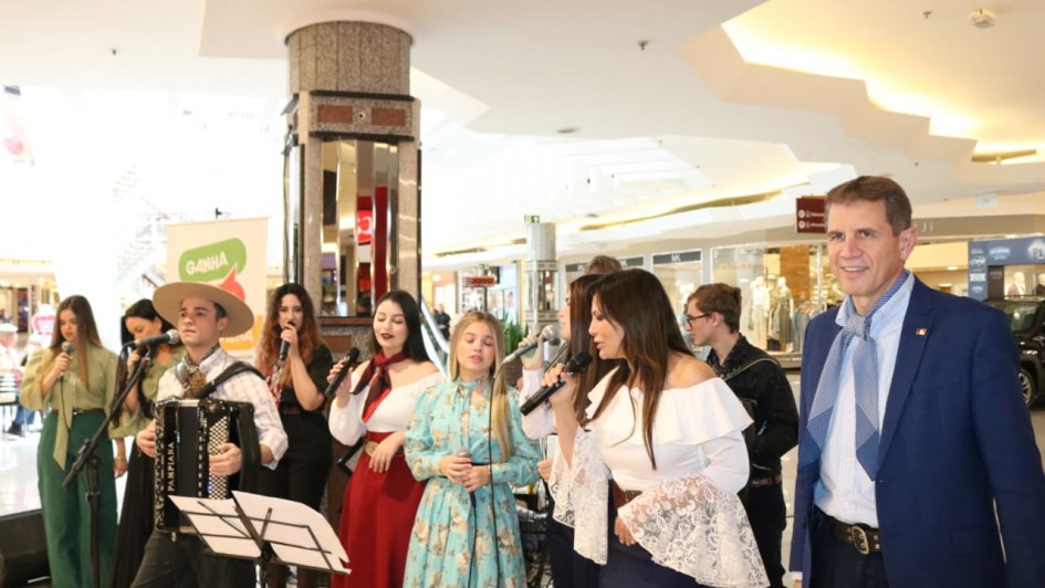 Um grupo de cantores e músicos nativistas se apresenta no shopping. Eles vestem roupas tradicionais gaúchas. No canto direito da imagem, está o secretário de desenvolvimento econômico, de lenço e bombacha.