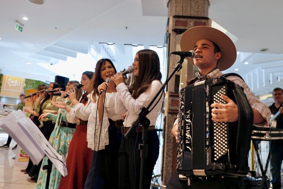 Diversos artistas nativistas estão cantando em um shopping. No canto direito, há um rapaz tocando gaita e cantando ao microfone. Eles vestem roupas tradicionais gaúchas.