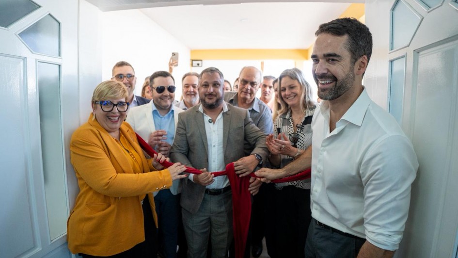 Um grupo de pessoas participa de uma cerimônia de inauguração em ambiente interno, de frente para uma porta branca aberta. Ao centro, uma mulher de blazer amarelo-mostarda e óculos de armação grossa azul segura uma tesoura grande e sorri, pronta para cortar a faixa vermelha que simboliza a abertura oficial. Do lado direito, um homem de camisa branca de mangas dobradas também segura a faixa e sorri amplamente. Atrás deles, várias pessoas, homens e mulheres, acompanham a cena, algumas batendo palmas e outras registrando o momento com celular. O clima é festivo e descontraído.