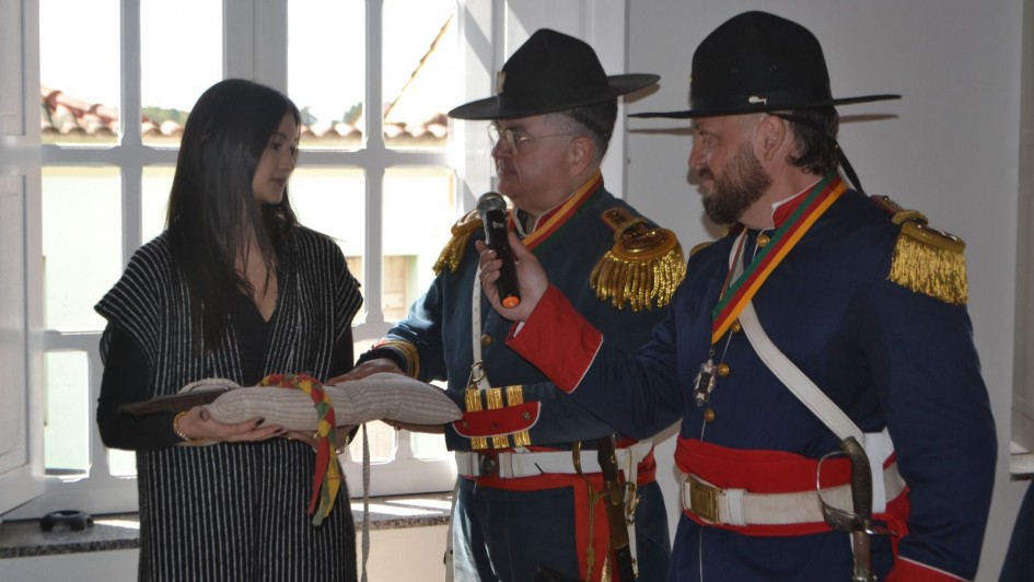 Dois homens vestidos com trajes históricos de inspiração militar farroupilha, incluindo fardas azuis com detalhes vermelhos, ombreiras douradas e chapéus de aba larga, participam de uma solenidade em ambiente interno. Um deles segura um microfone enquanto o outro observa. À esquerda, uma mulher de cabelos longos e escuros veste roupa preta com sobreposição listrada e segura uma almofada com uma lança histórica apoiada, envolta em uma faixa colorida nas cores do Rio Grande do Sul. Ao fundo, há uma janela ampla iluminando a cena.