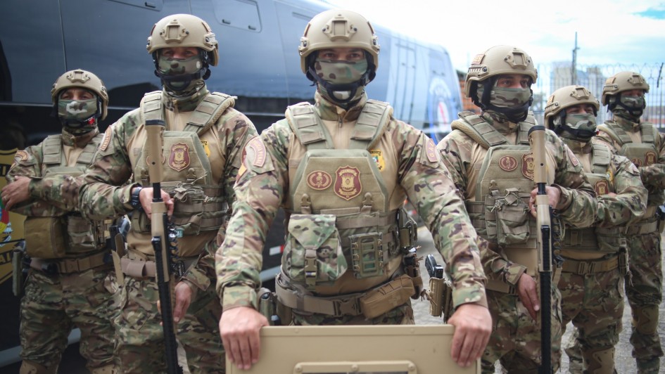 Grupo de agentes do Grupo de Ações Especiais da Polícia Penal (GAES) em formação, vestidos com uniformes camuflados completos, incluindo capacetes, coletes táticos e máscaras. O agente central segura um escudo de proteção, enquanto outros portam armas longas. Ao fundo, há um ônibus preto da Polícia Penal.