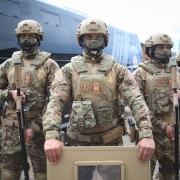 Grupo de agentes do Grupo de Ações Especiais da Polícia Penal (GAES) em formação, vestidos com uniformes camuflados completos, incluindo capacetes, coletes táticos e máscaras. O agente central segura um escudo de proteção, enquanto outros portam armas longas. Ao fundo, há um ônibus preto da Polícia Penal.