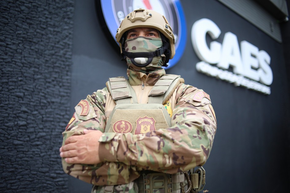 Agente do GAES em destaque, em uniforme camuflado e capacete, com os braços cruzados. Ele usa máscara que cobre parte do rosto e colete com brasões da Polícia Penal. Ao fundo, aparece a parede escura com o letreiro “GAES”.
