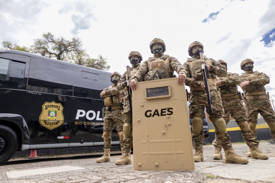 Linha de agentes do GAES posicionados em frente a um ônibus preto da Polícia Penal. No centro, um policial segura um escudo bege com a inscrição “GAES”. Todos usam uniformes camuflados, capacetes e equipamentos de proteção.