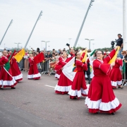 Mulheres dançando na rua com vestidos vermelhos e bandeiras coloridas. Sorriem enquanto o público assiste atrás de notas. É um momento alegre e cheio de movimento.