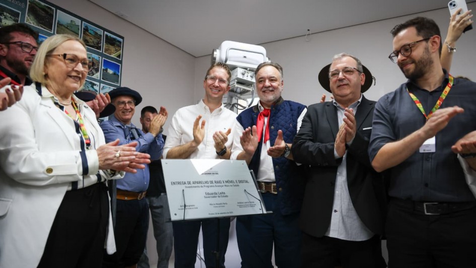 A imagem mostra um evento de inauguração de um aparelho de raio-X em Piratini. Vários indivíduos estão reunidos, aplaudindo. No centro da imagem, um grupo de pessoas está em destaque, posando ao lado de uma placa de metal com inscrições, provavelmente detalhando a entrega do equipamento. Entre os presentes, alguns estão vestindo trajes típicos, como o chapéu e o lenço vermelho, indicando uma referência cultural, possivelmente gaucha. A pessoa à esquerda usa óculos e um casaco branco, enquanto os outros estão usando diferentes trajes formais e casuais. Eles parecem celebrar a entrega do aparelho, que é visível ao fundo.
