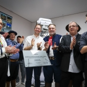 A imagem mostra um evento de inauguração de um aparelho de raio-X em Piratini. Vários indivíduos estão reunidos, aplaudindo. No centro da imagem, um grupo de pessoas está em destaque, posando ao lado de uma placa de metal com inscrições, provavelmente detalhando a entrega do equipamento. Entre os presentes, alguns estão vestindo trajes típicos, como o chapéu e o lenço vermelho, indicando uma referência cultural, possivelmente gaucha. A pessoa à esquerda usa óculos e um casaco branco, enquanto os outros estão usando diferentes trajes formais e casuais. Eles parecem celebrar a entrega do aparelho, que é visível ao fundo.