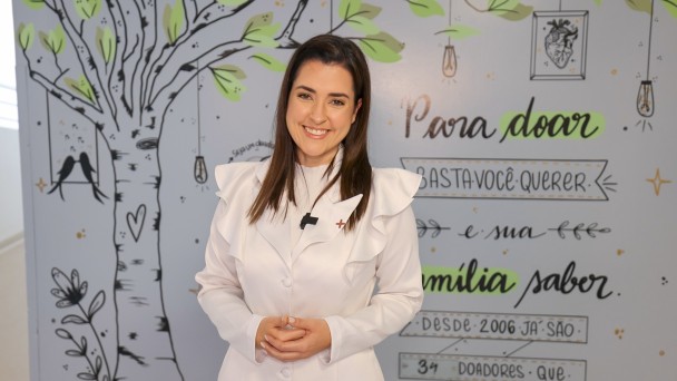 Uma mulher vestida de branco está em pé, sorrindo para a câmera com as mãos cruzadas. Ao fundo, há um painel com desenho de uma árvore, com flores, folhas, corações e luzes, onde está escrito: "Para doar basta você querer e sua família saber". 