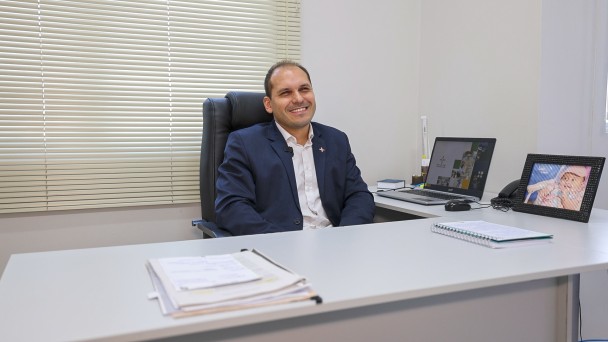 Um homem vestindo roupas sociais está sentado em sua mesa de trabalho, sorrindo para a foto. Sobre a mesa, há um notebook, um porta-retrato com a imagem de um nenê e cadernos e folhas com anotações. 