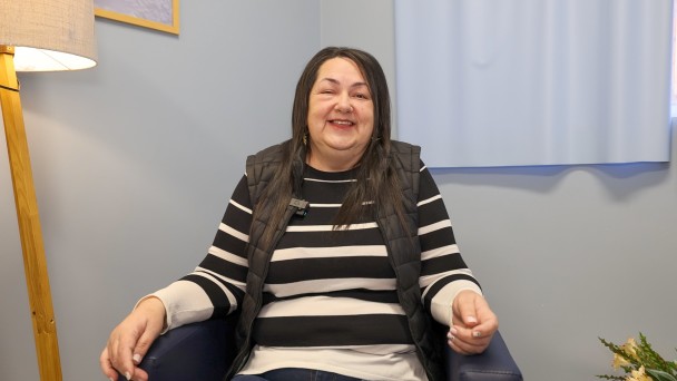 Uma mulher de cabelos escuros e pele branca está sentada em uma poltrona, sorrindo para a foto. Ao lado, há uma luminária. 