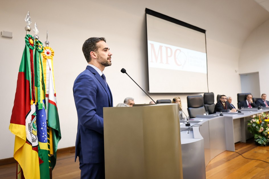 Leite, de terno azul, aparece em destaque, falando ao microfone no púlpito. Atrás dele estão as bandeiras e, ao fundo, uma tela de projeção com a sigla “MPC”. À mesa, várias pessoas de terno e toga acompanham a cerimônia.