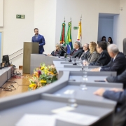 Leite está de terno azul e em pé, falando em um púlpito em um auditório. Na mesa em frente a ele, várias pessoas estão sentadas em cadeiras altas, ouvindo a fala. Há bandeiras do Brasil e do Rio Grande do Sul ao fundo, além de arranjos de flores na frente da mesa.