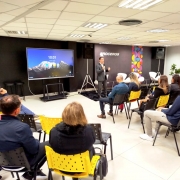 Na imagem, um homem em traje formal fala em pé diante de um grupo de pessoas sentadas em cadeiras pretas e amarelas organizadas em fileiras. Ele está posicionado próximo a um telão. O ambiente é uma sala moderna, bem iluminada, com teto de placas brancas, paredes escuras e o logotipo “PROCERGS” em destaque. 