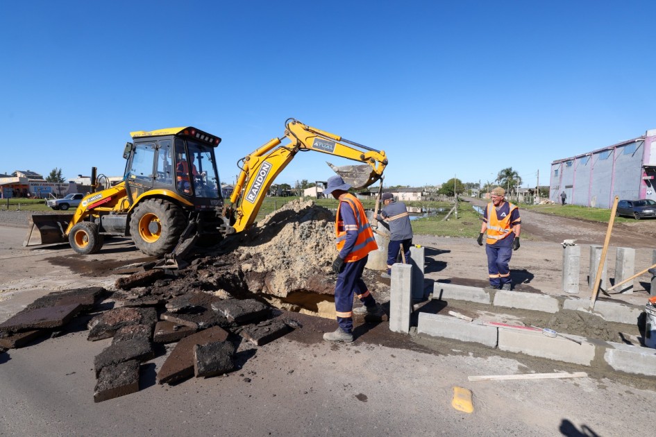 Operários com coletes laranja trabalham em uma obra viária durante o dia. Uma retroescavadeira amarela retira terra e pedaços de asfalto quebrado. No entorno, há blocos de concreto sendo assentados e ferramentas espalhadas. O cenário mostra uma área urbana simples, com algumas casas e prédios ao fundo.