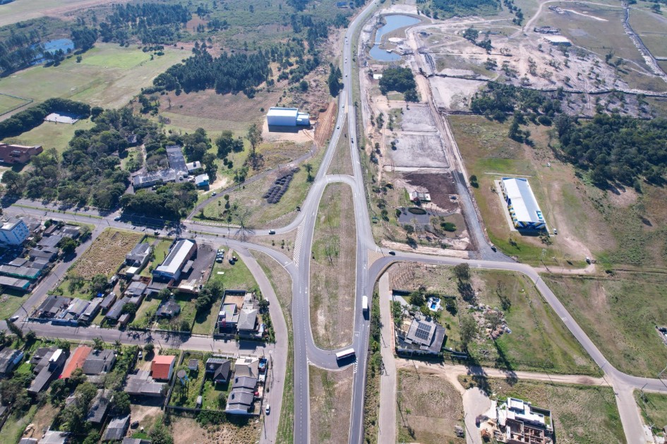 Imagem aérea mostra uma rodovia duplicada com canteiro central e alças de acesso, cercada por áreas urbanas e rurais. Há residências, galpões e áreas verdes ao redor. A estrada tem tráfego leve de veículos, destacando a organização do novo entroncamento viário.